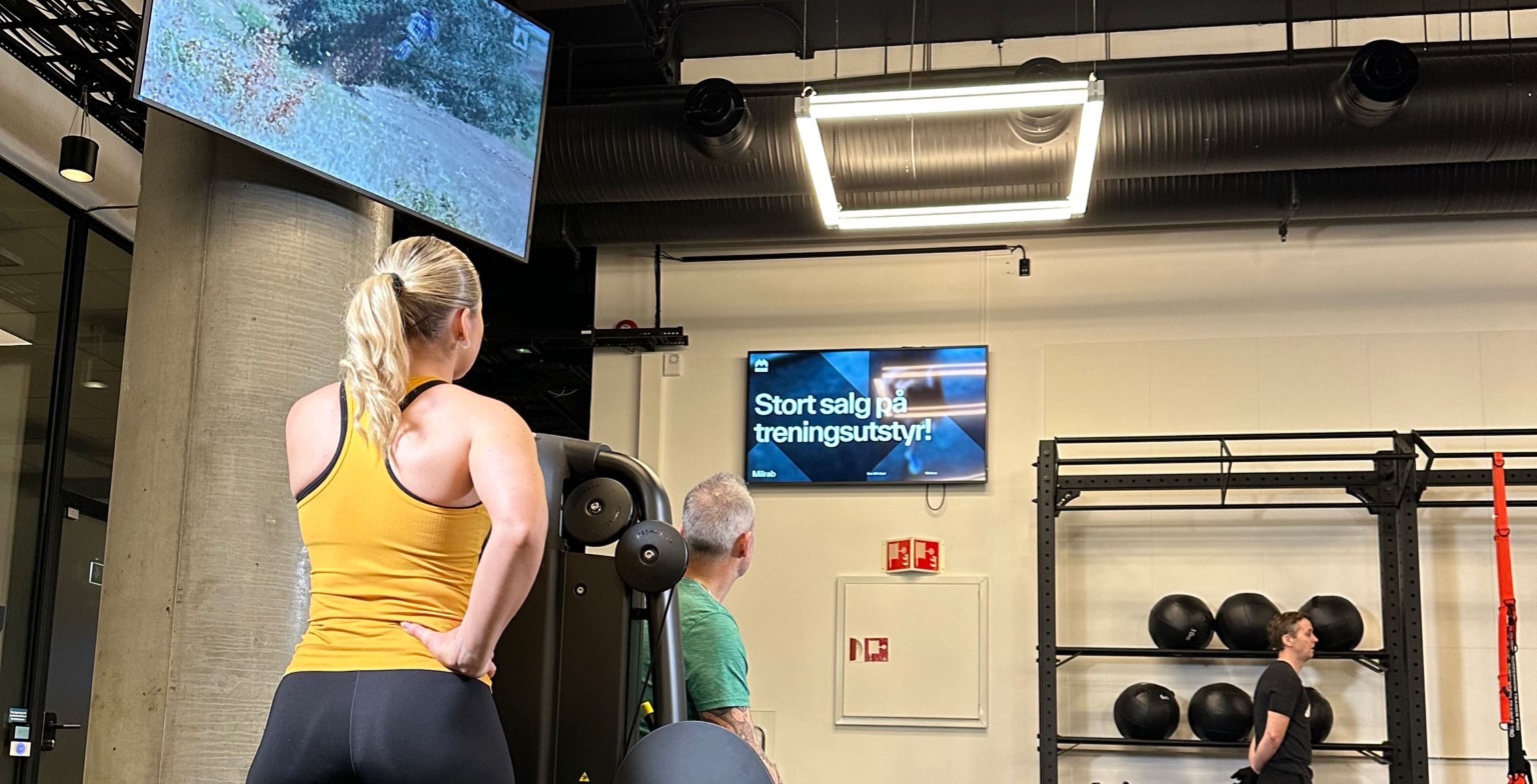 Kvinne i gul treningstopp ser opp mot en skjerm i taket. På veggen bak viser en annen skjerm en annonse for treningsutstyr.